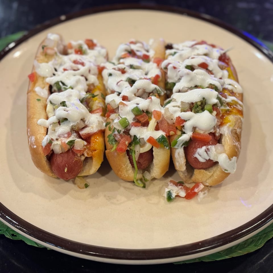 Three hot dogs in buns, topped with pico de gallo, cheese, and a creamy white sauce.
