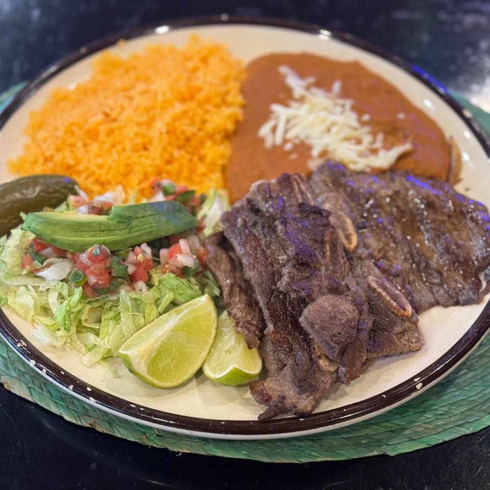 Grilled short ribs with rice, refried beans, salad, avocado, lime, and a pickle.