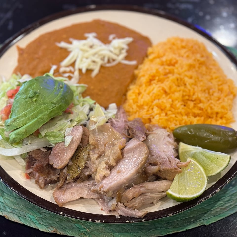 Mexican carnitas plate with rice, refried beans, avocado, lettuce, lime, and a jalapeño.