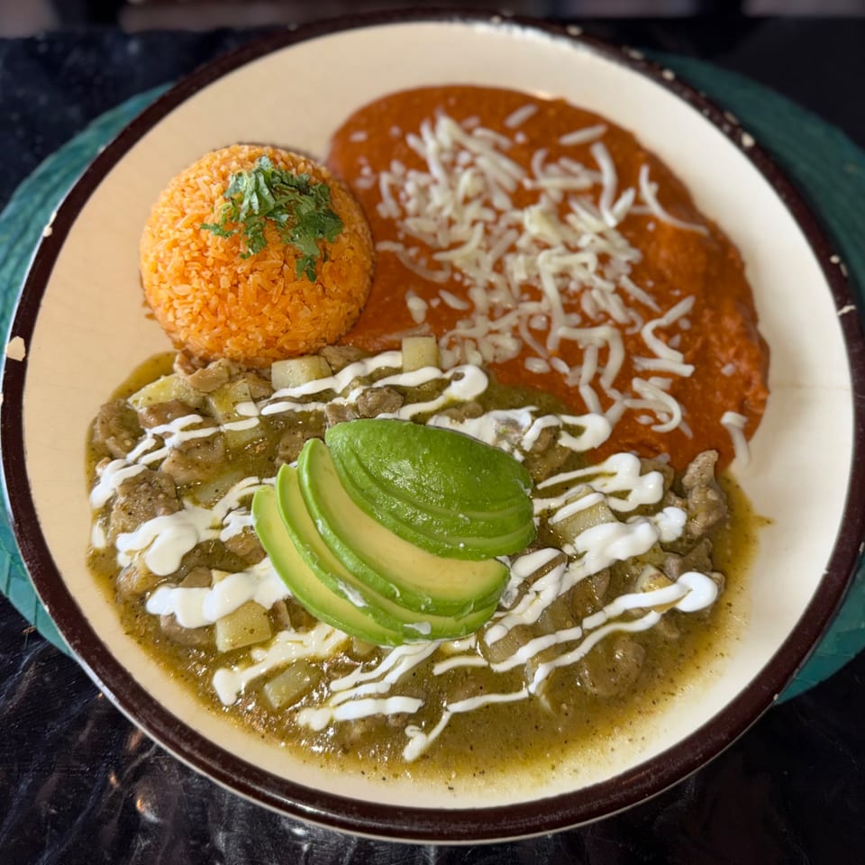 A plate of Mexican food with red rice, green chile stew, red enchilada sauce, and avocado slices.