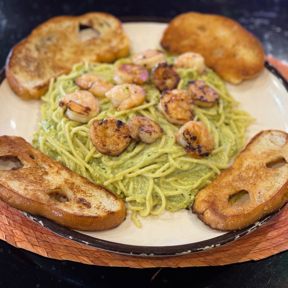 Green pesto pasta with grilled shrimp and toasted bread on a white plate.