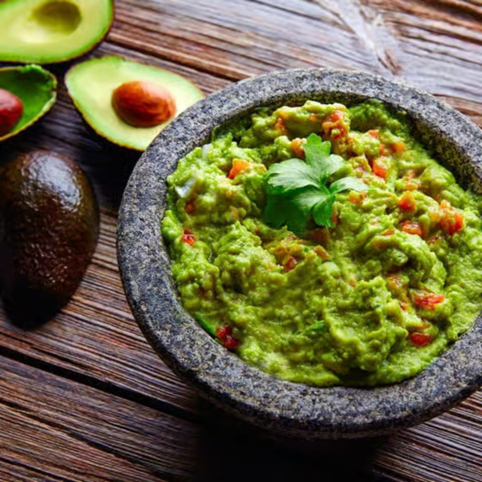 Fresh guacamole in a stone molcajete, garnished with cilantro, alongside whole and halved avocados.