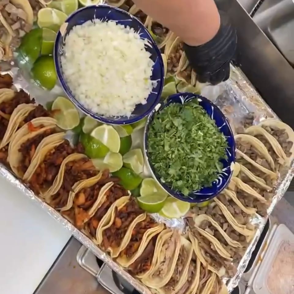 A tray of various tacos with bowls of diced onions, cilantro, and lime wedges.
