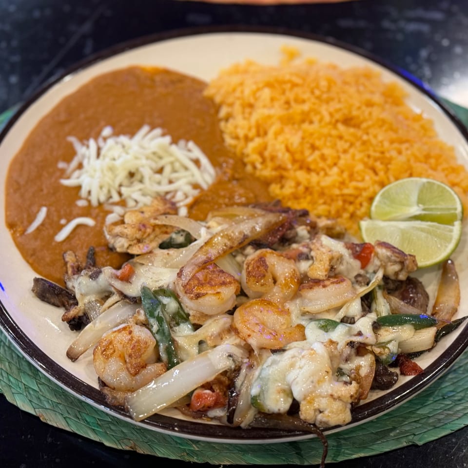 A plate of shrimp fajitas with onions, peppers, rice, refried beans, and lime wedges.