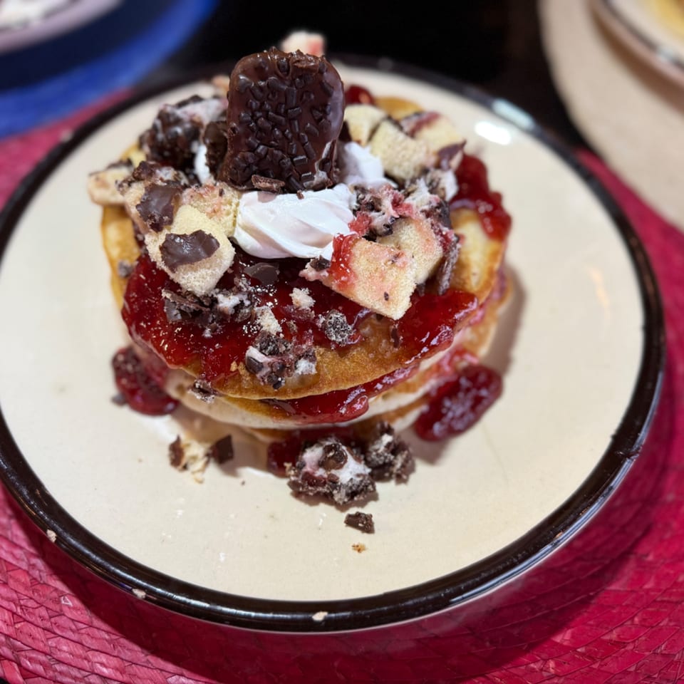 Stack of pancakes with strawberry jam, whipped cream, chocolate chunks, and a chocolate bar.