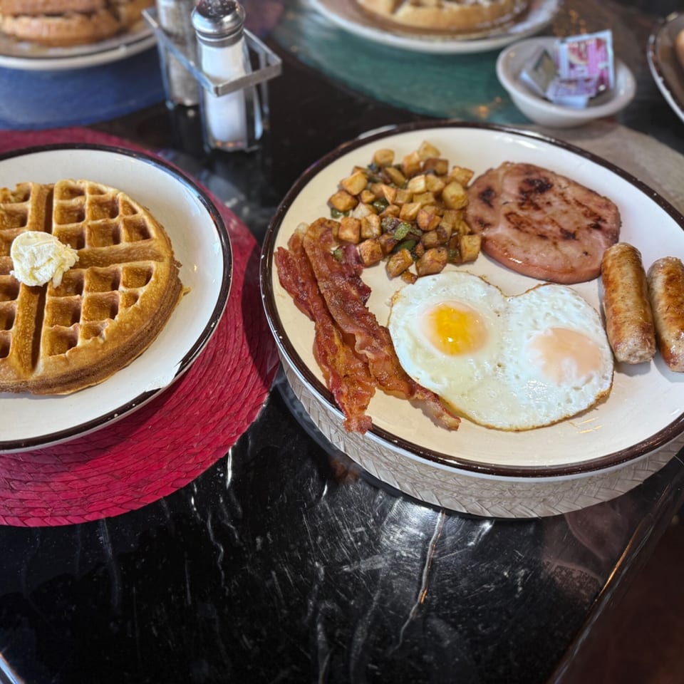 A full breakfast plate with fried eggs, bacon, sausage, ham, and home fries next to a waffle.