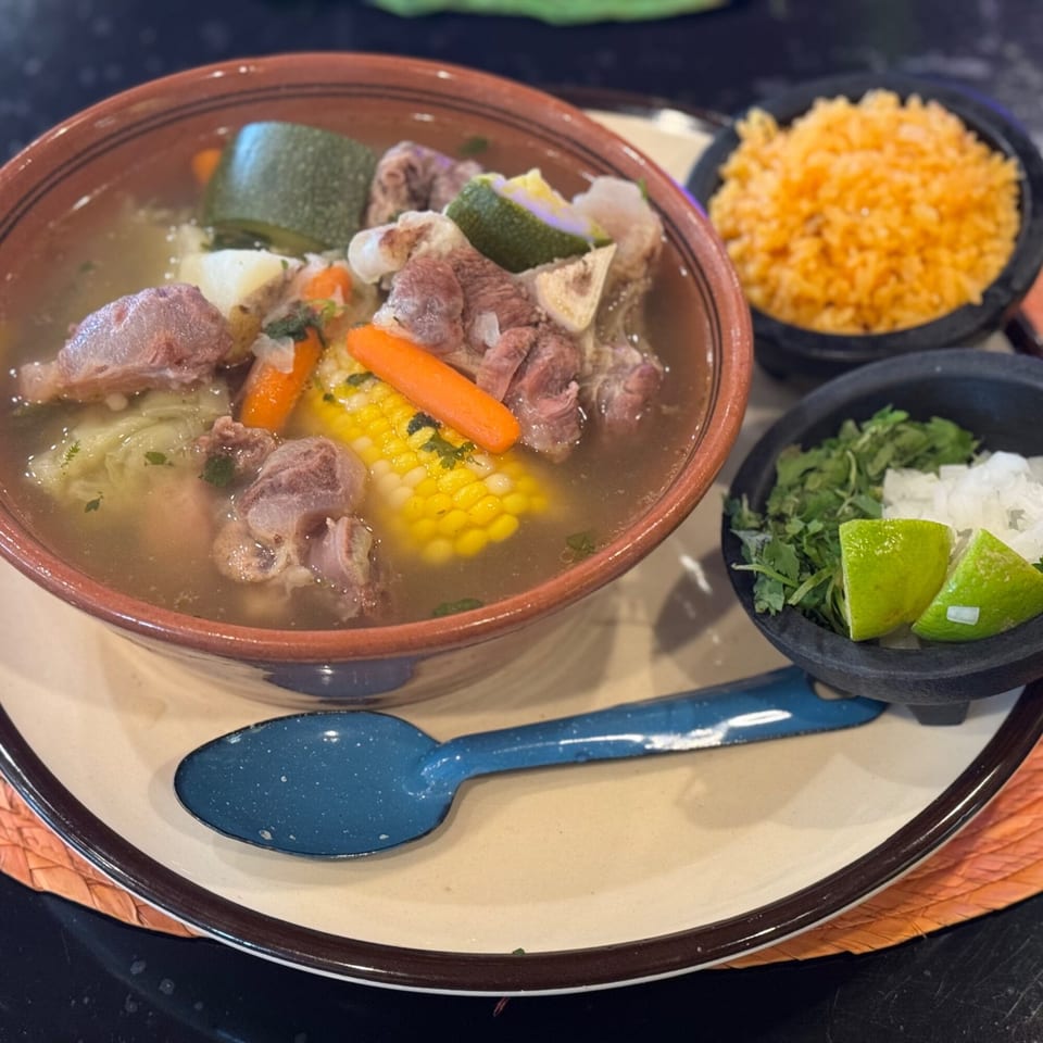 Hearty beef soup with corn, carrots, and zucchini, served with rice, cilantro, onions, and lime.