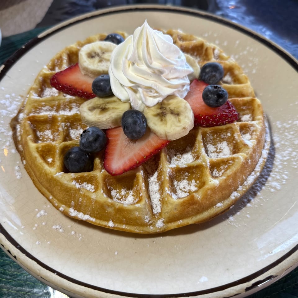Golden waffle topped with whipped cream, sliced bananas, strawberries, and blueberries, dusted with powdered sugar.