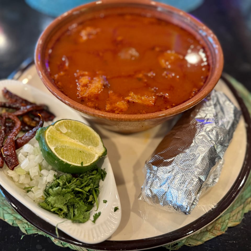 A bowl of red stew with a side of dried chiles, lime, chopped onions, cilantro, and a foil-wrapped item.